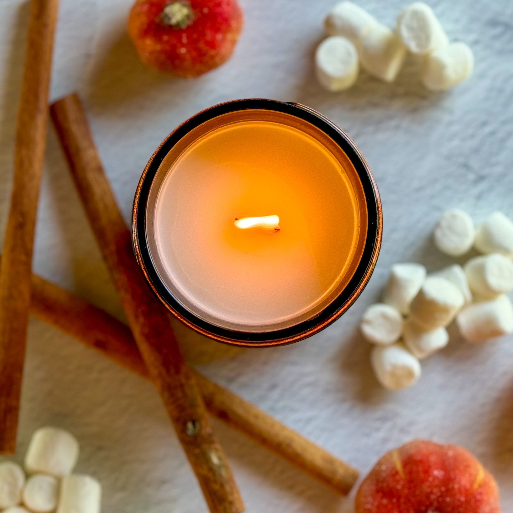 Overhead view of lit candle surrounded by cinnamon sticks, small pumpkins, and marshmallows on a light background
