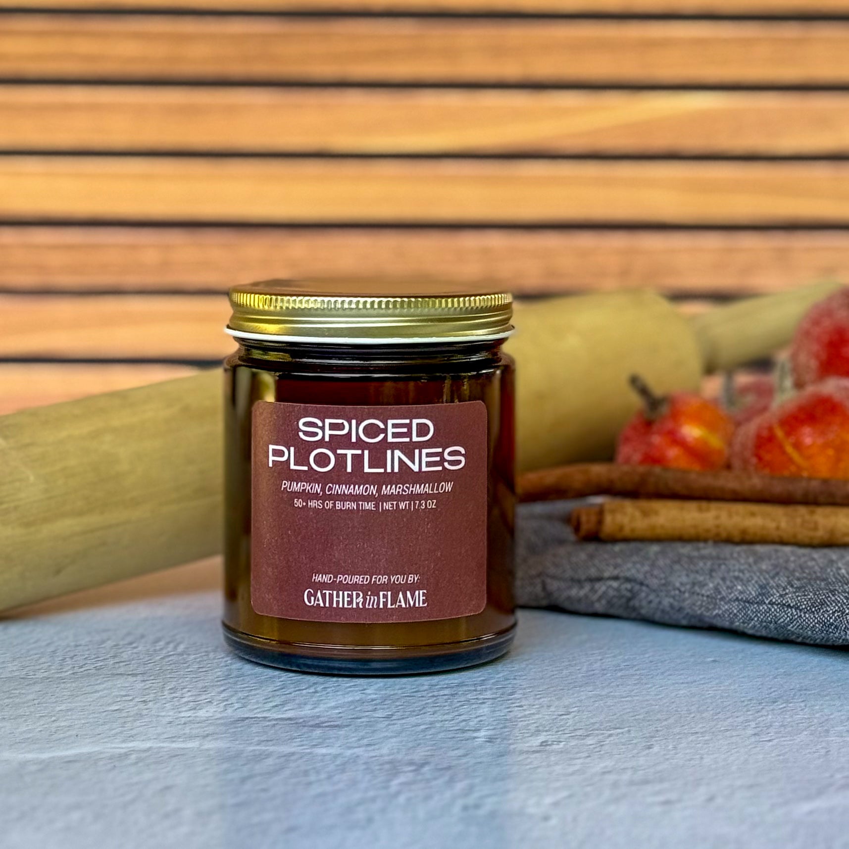 Jar of 'Spiced Plotlines' candle with cinnamon sticks, a rolling pin and pumpkins in the background
