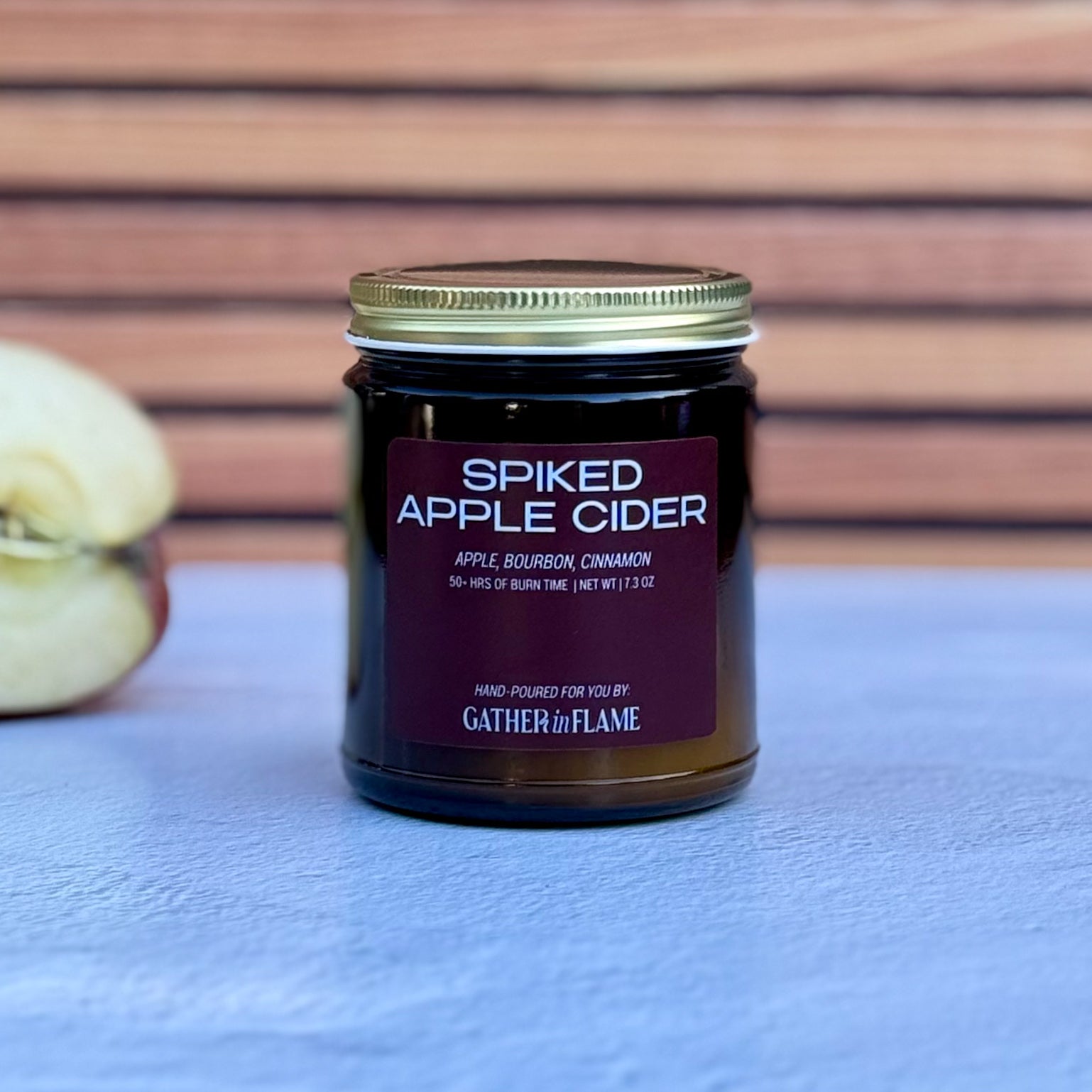 Jar of 'Spiked Apple Cider' candle with a purple label on a white surface with apple in the background