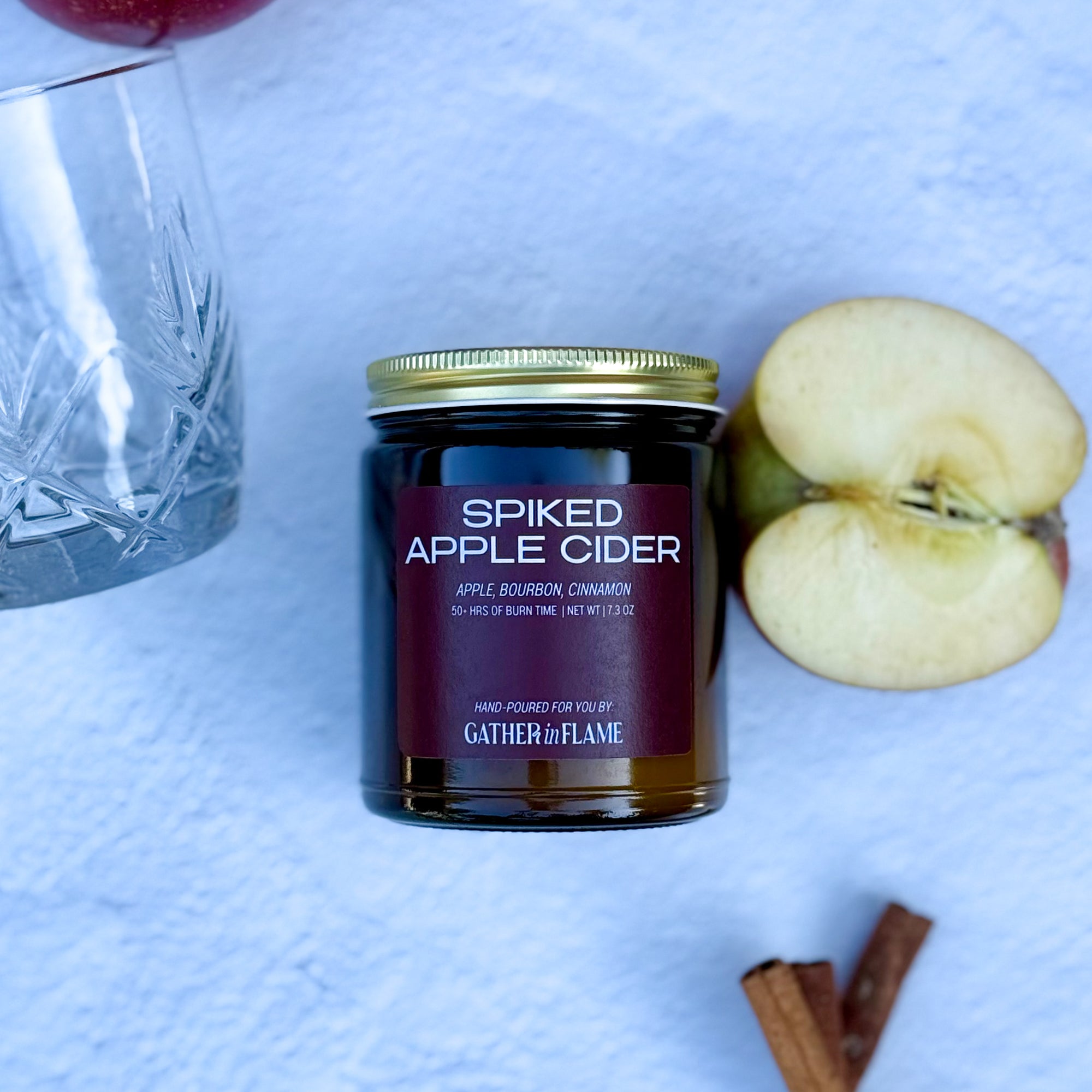 Flatlay of 'Spiked Apple Cider' candle with apples, a whiskey glass and cinnamon sticks on a white background