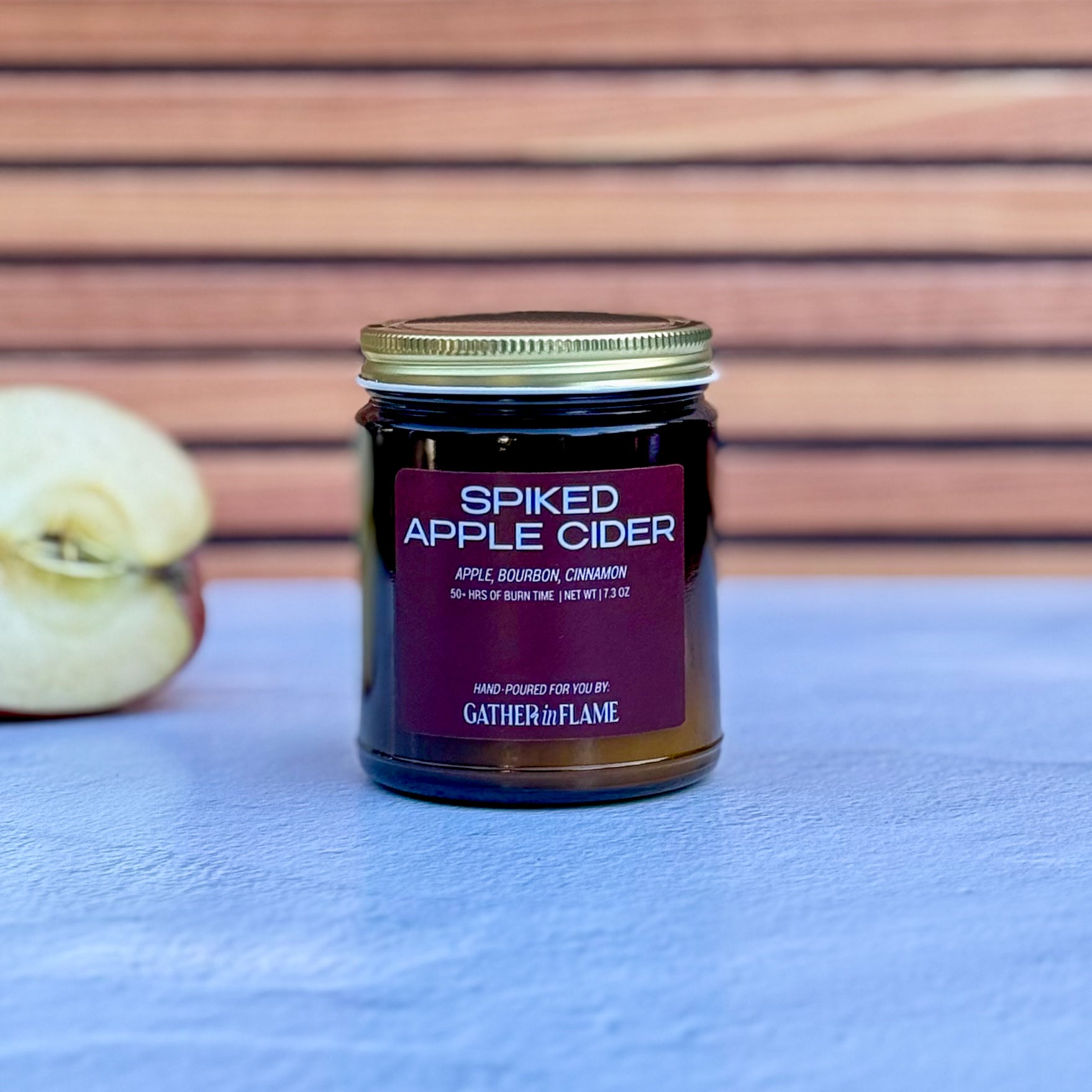 Jar of 'Spiked Apple Cider' candle with an apple on a blue surface and wooden background