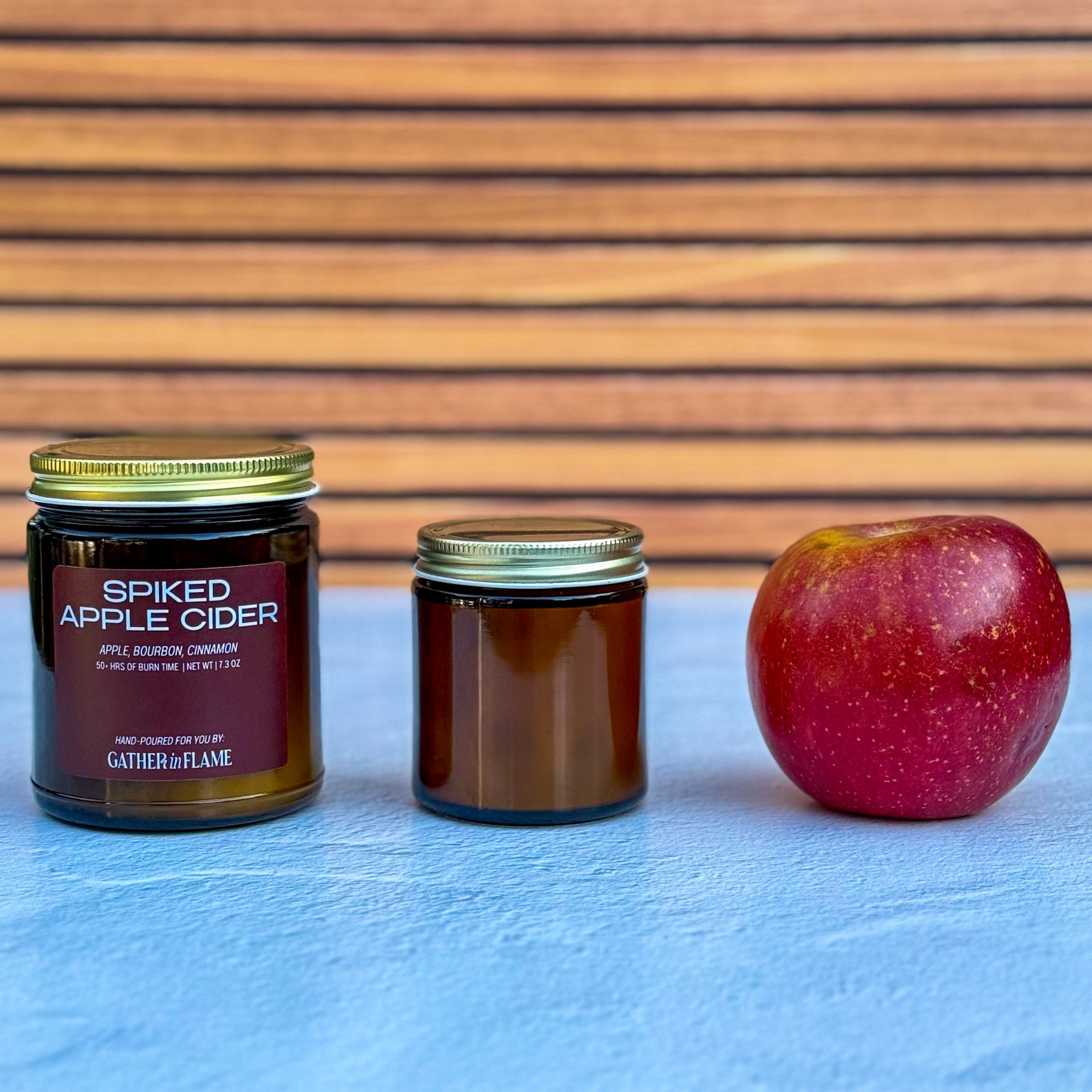Two differently sized candles labeled &#39;Spiked Apple Cider&#39; and a red apple on a blue surface with a wooden background.