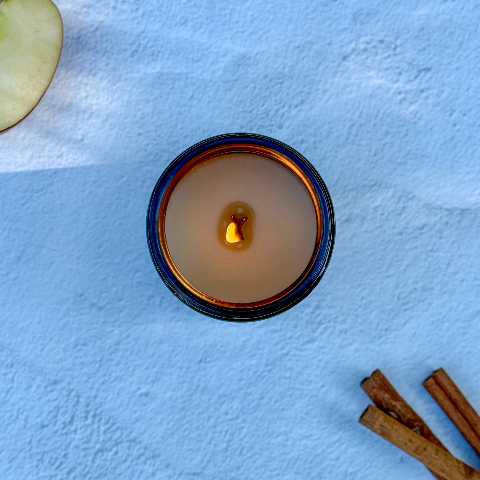 Overhead photo of a lit candle surface with wood wick with apple and cinnamon sticks on light background