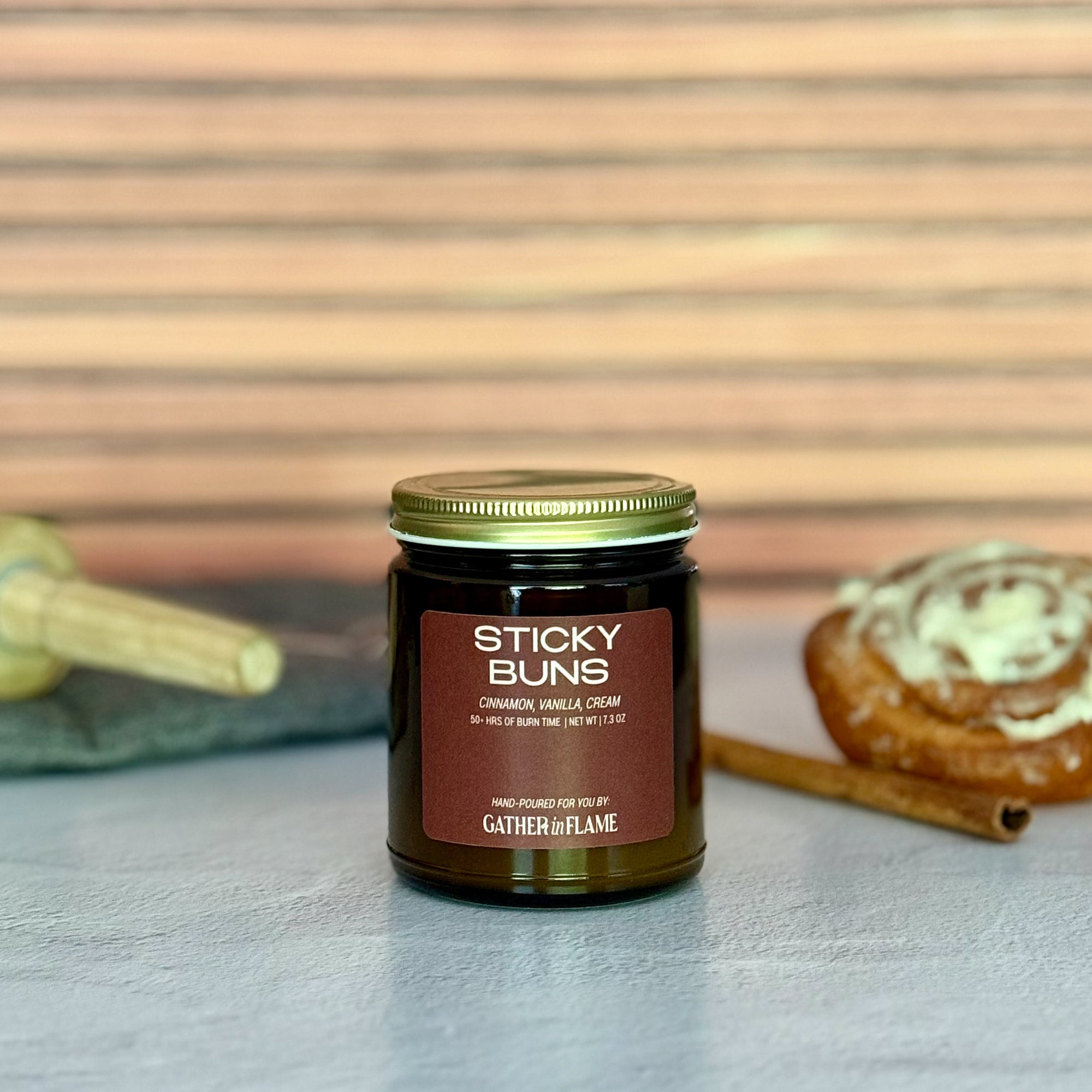 Jar of 'Sticky Buns' candle with a cinnamon roll, rolling pin and cinnamon stick on a wooden surface.