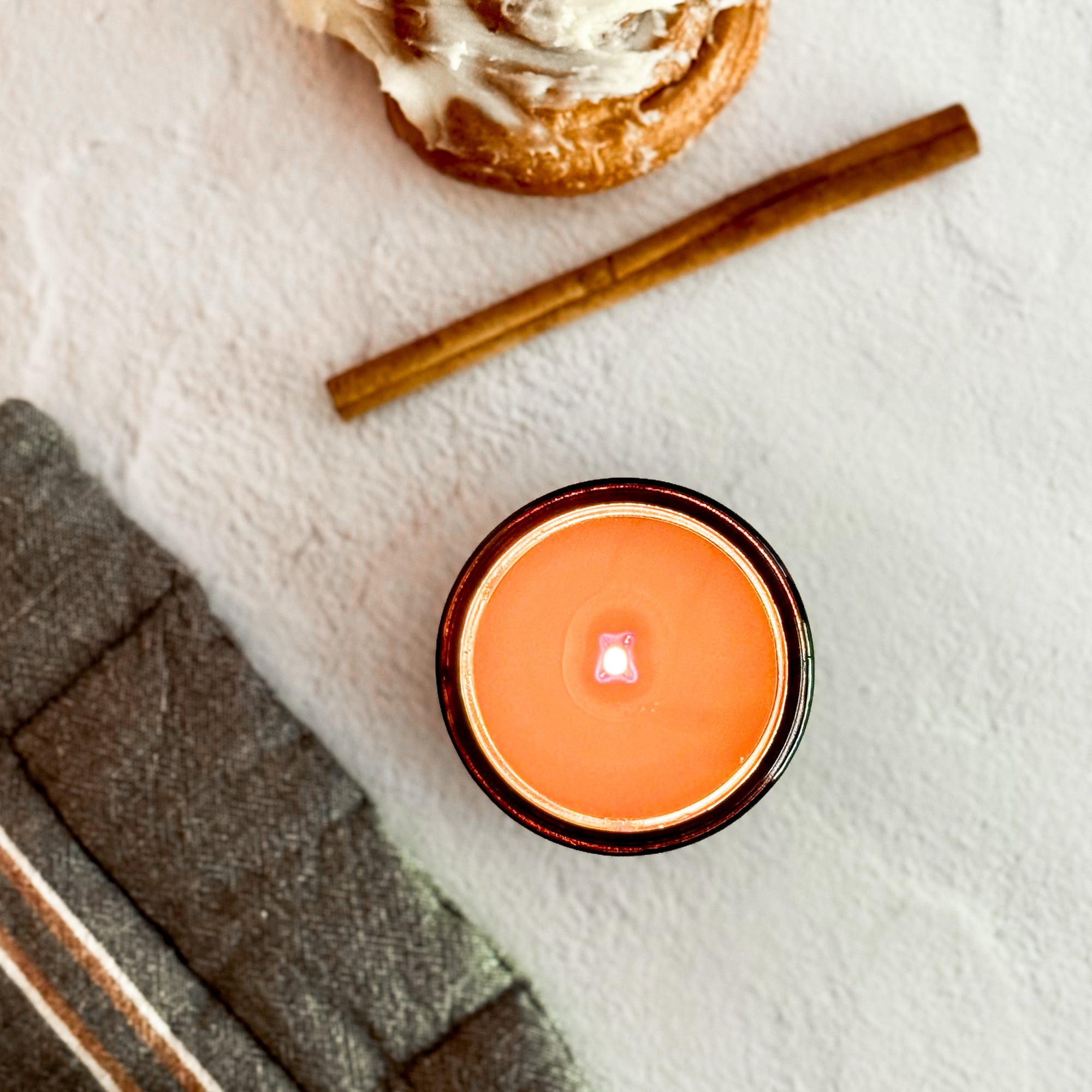 Overhead view of lit candle on a textured surface with cinnamon stick and cinnamon roll.