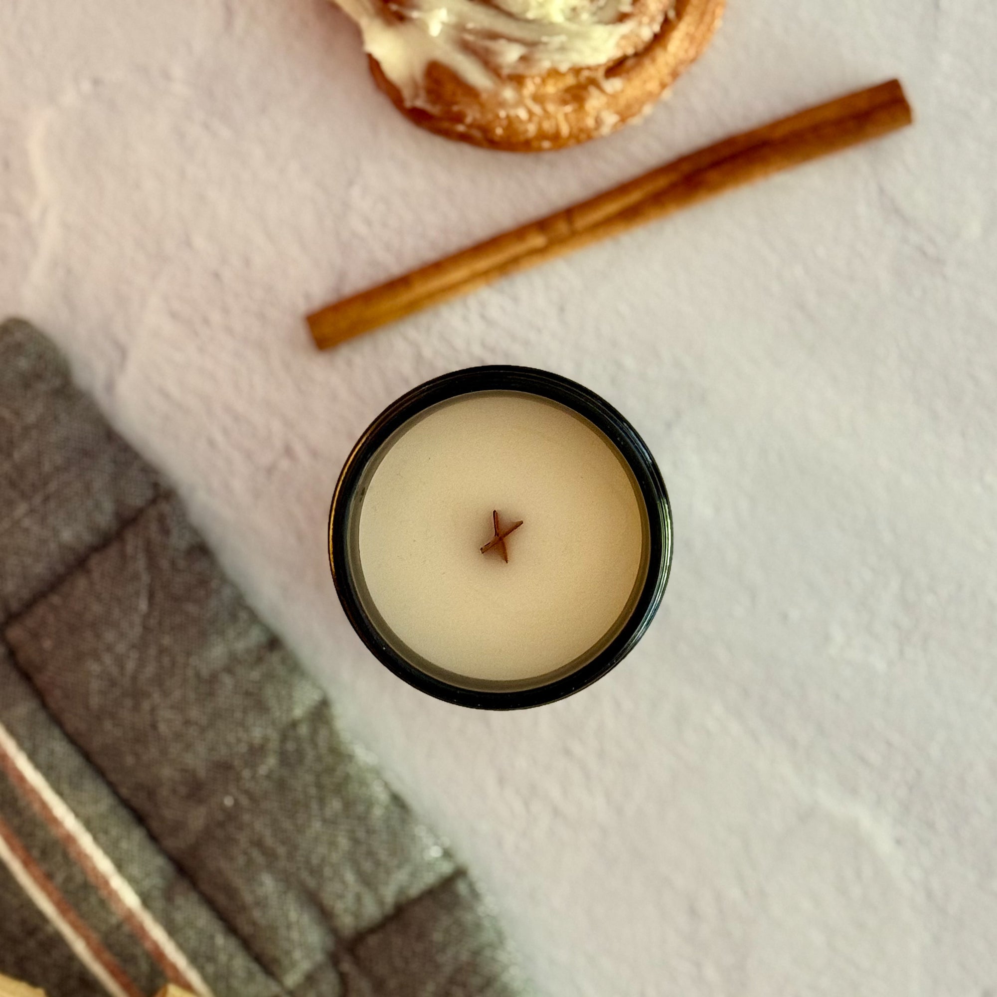 Overhead view of candle with wood wick on a textured surface with cinnamon stick and cinnamon roll.