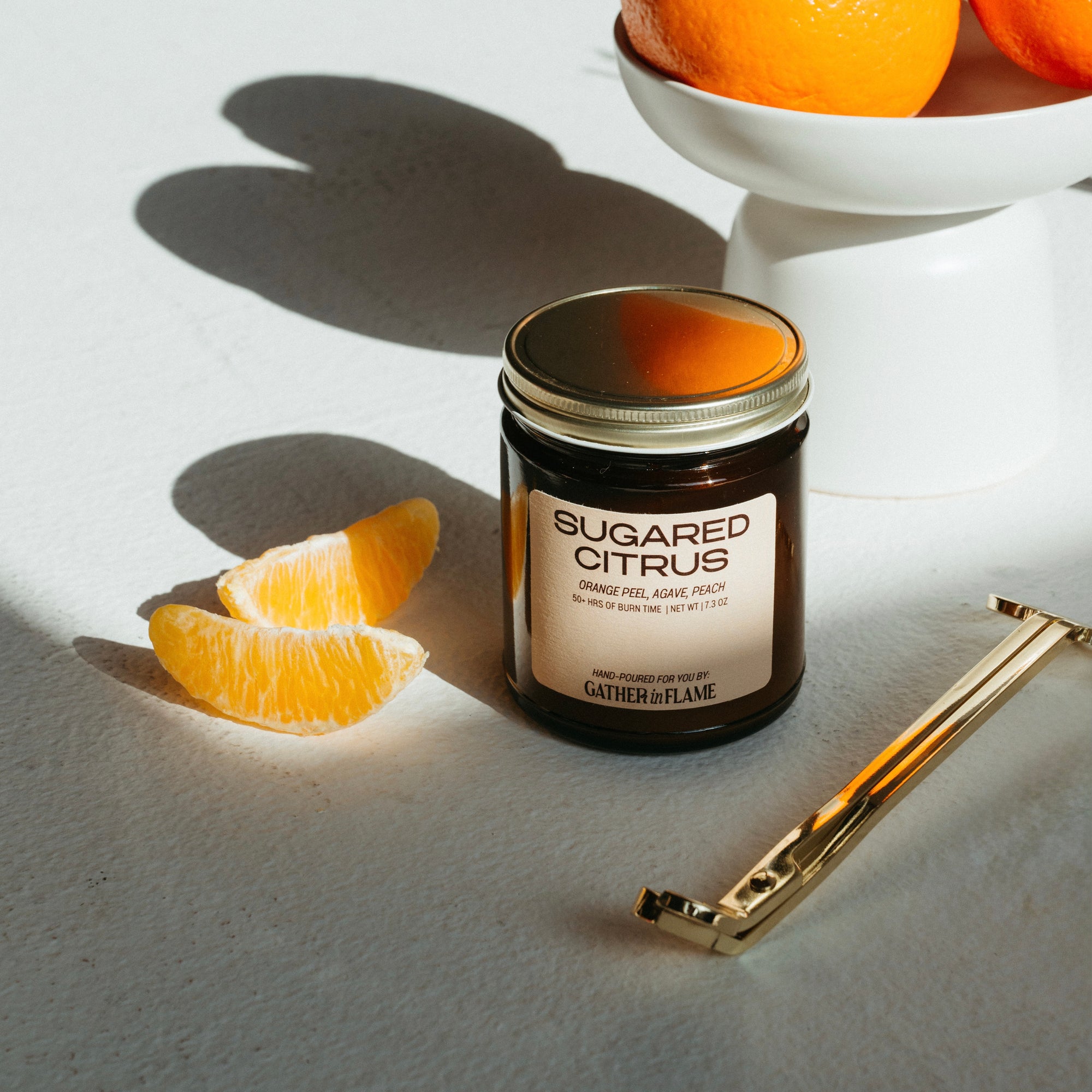 Jar of 'Sugared Citrus' candle with orange slices and a gold lighter on a light surface.