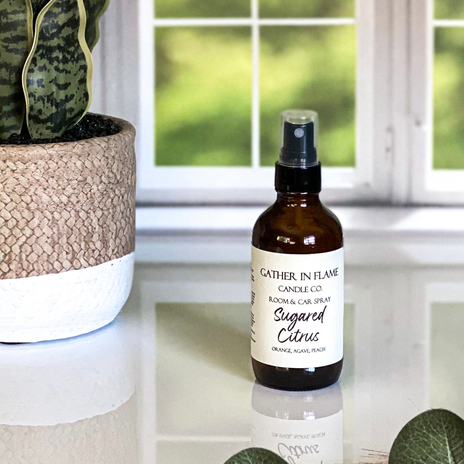 A brown spray bottle labeled 'GATHER IN FLAME CANDLE CO. ROOM & CAR SPRAY Sugared Citrus' placed on a white marble surface with a window and plant in the background.