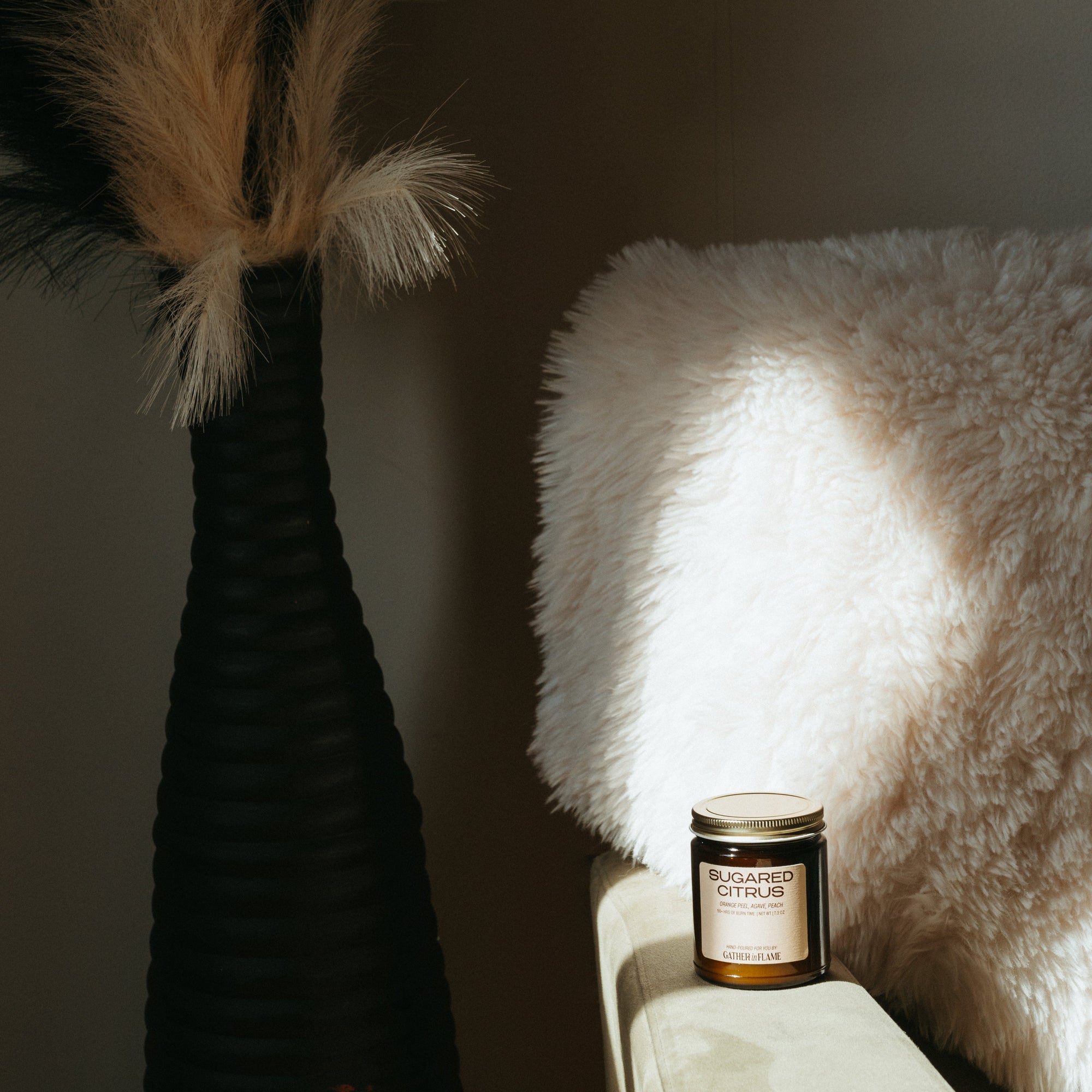 Candle on a surface next to a vase with pampas grass against a dark wall.
