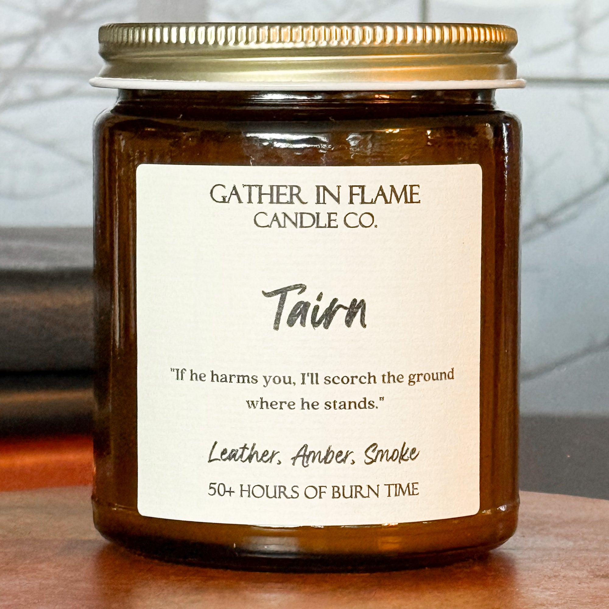 Close up of a jar candle labeled 'Tairn' by Gather in Flame Candle Co., placed on a wooden pedestal, with a window and books in the background.