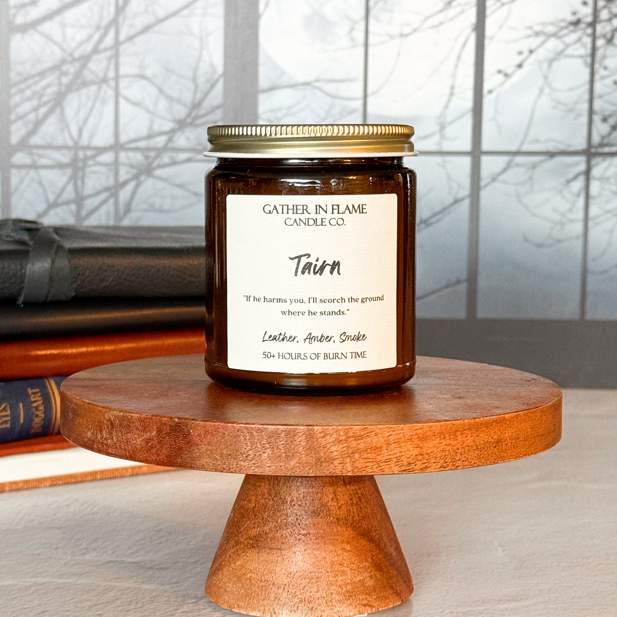A jar candle labeled 'Tairn' by Gather in Flame Candle Co., placed on a wooden pedestal, with a window and books in the background.