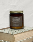 Amber glass candle Labeled The Archives on a stack of books with a white background.