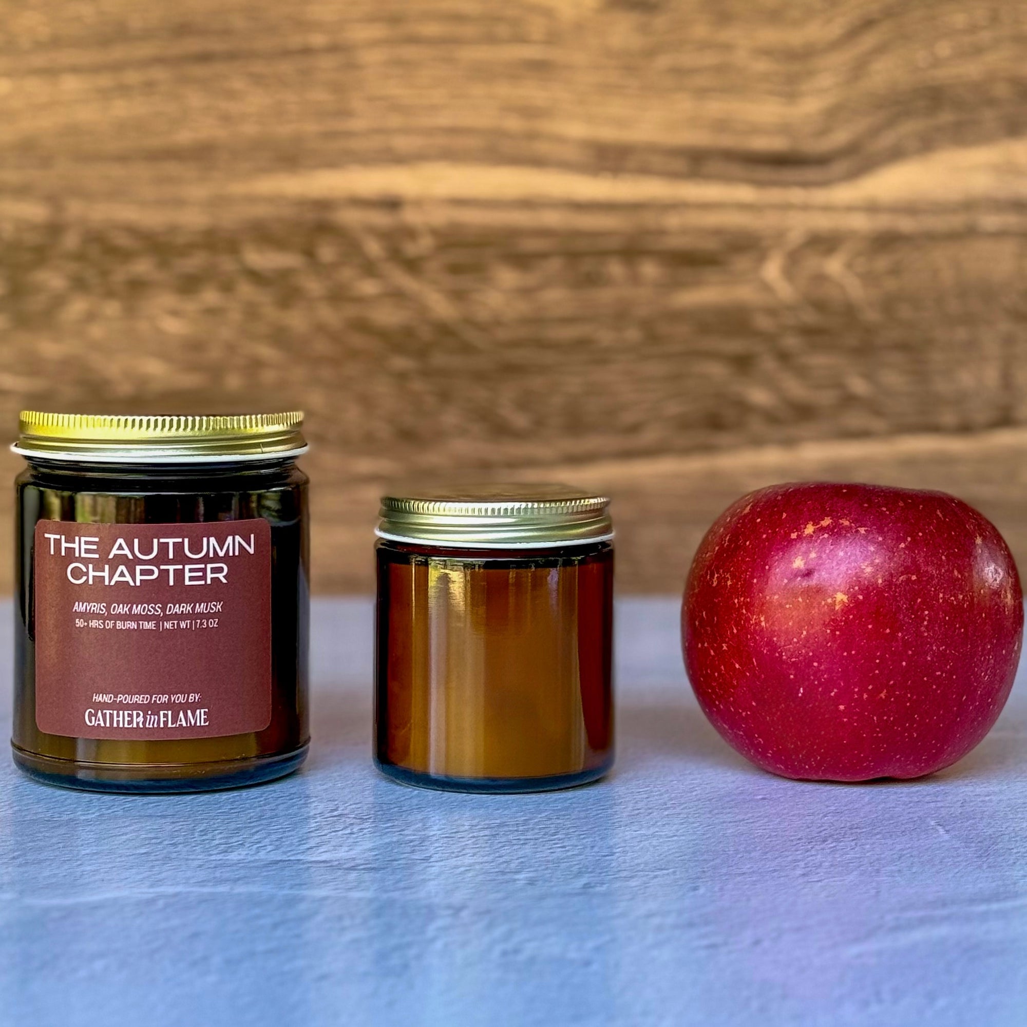Two candles of different sizes labeled The Autumn Chapter with apple on wooden background