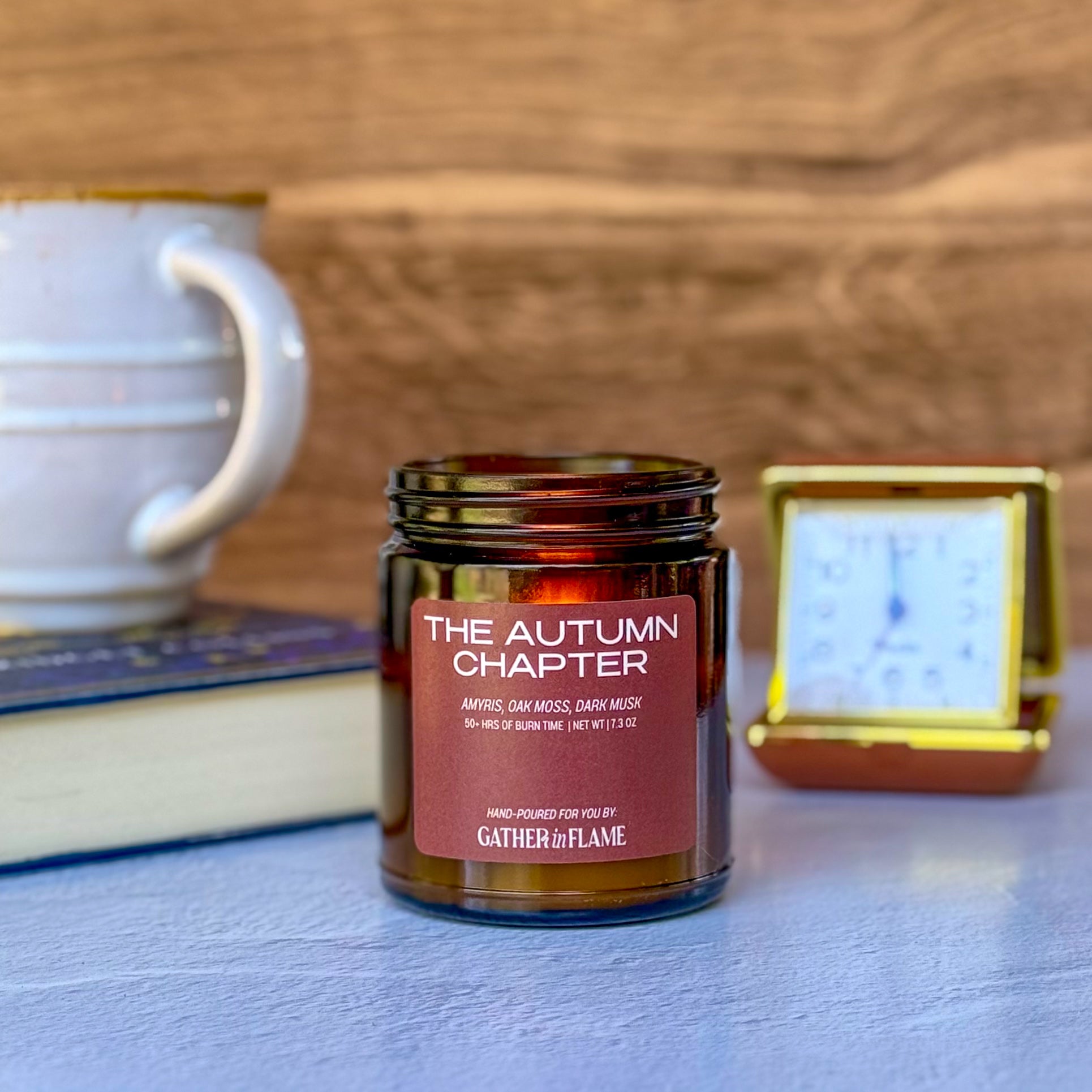 Lit candle labeled The Autumn Chapter with blurred background of book, mug and clock