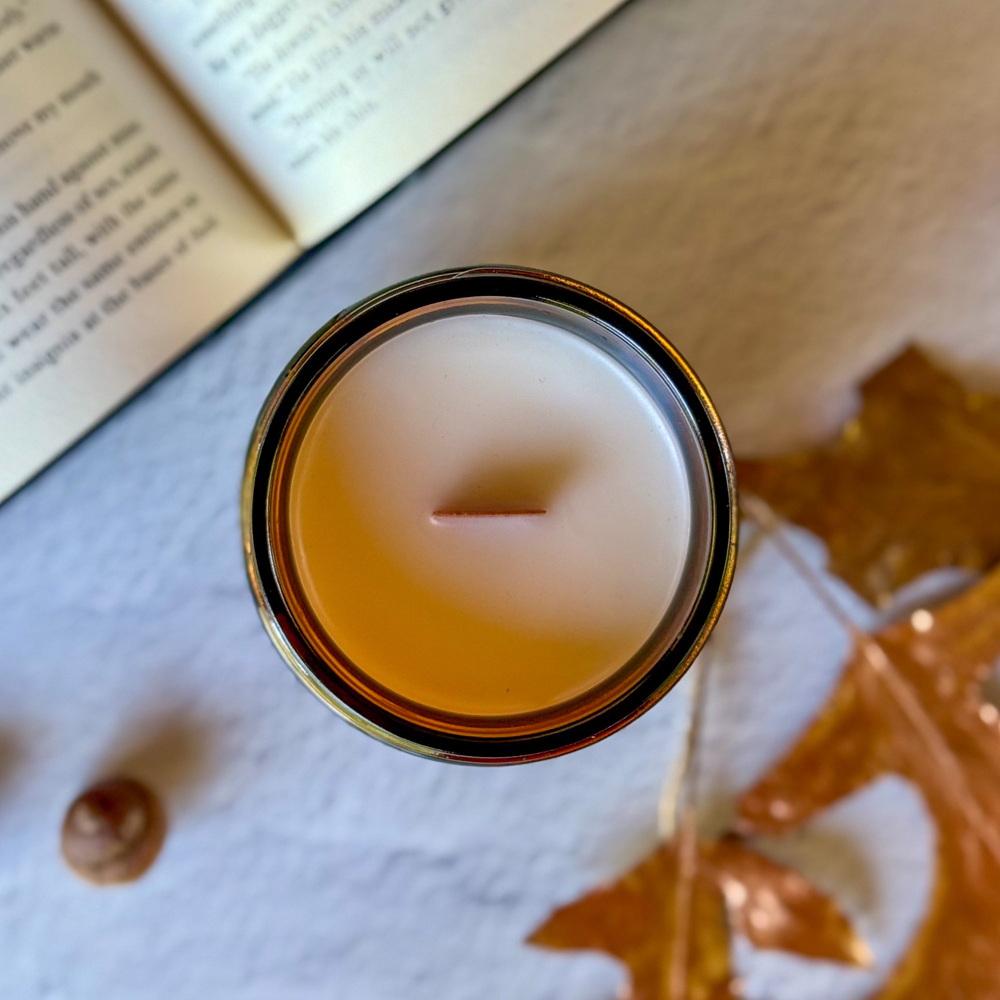 Overhead view of candle with wooden wick surrounded by blurred book, leaves and acorns