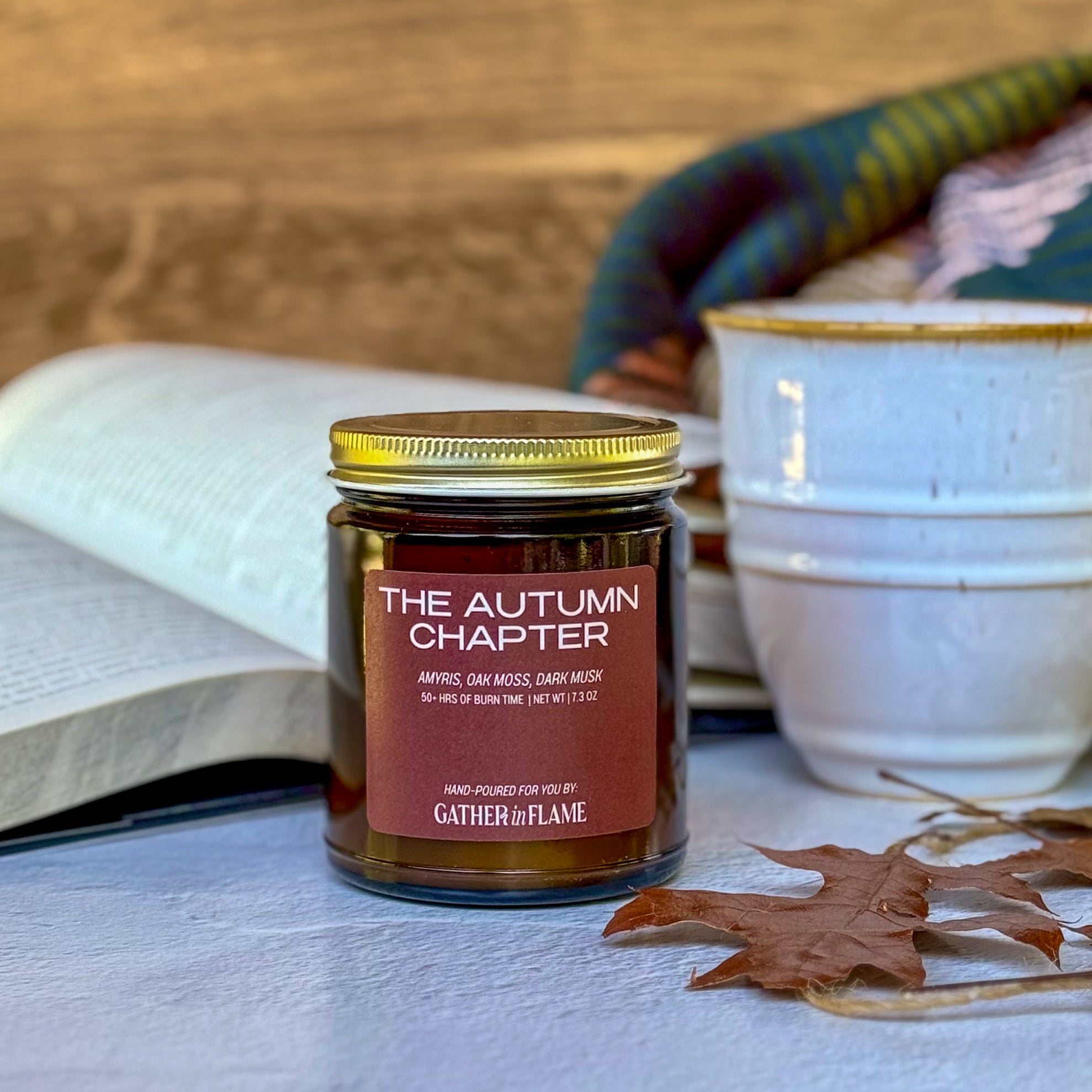 Candle labeled The Autumn Chapter with background of book, mug and plaid blanket