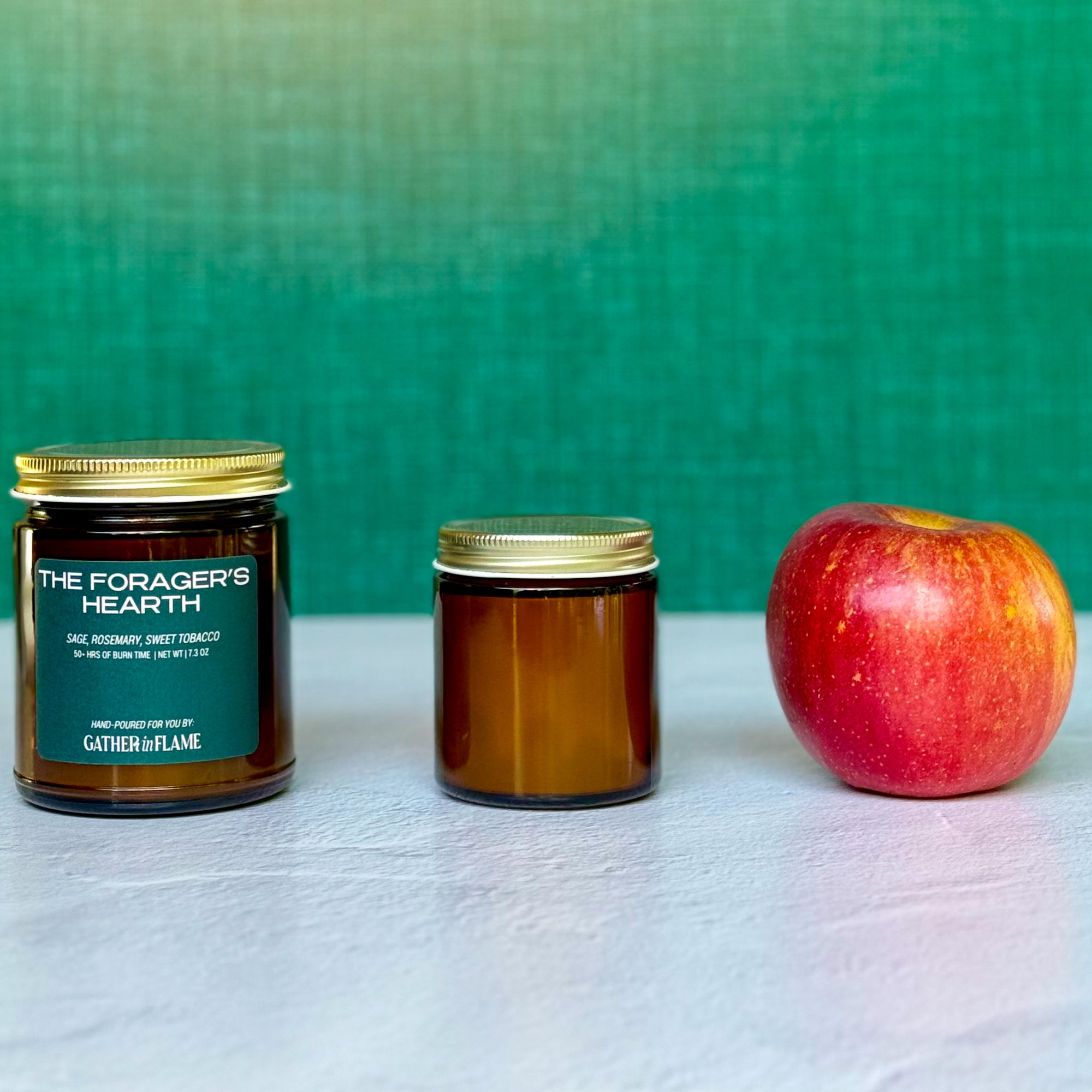 Two candles of different sizes labeled The Forager's Hearth next to an apple with green background