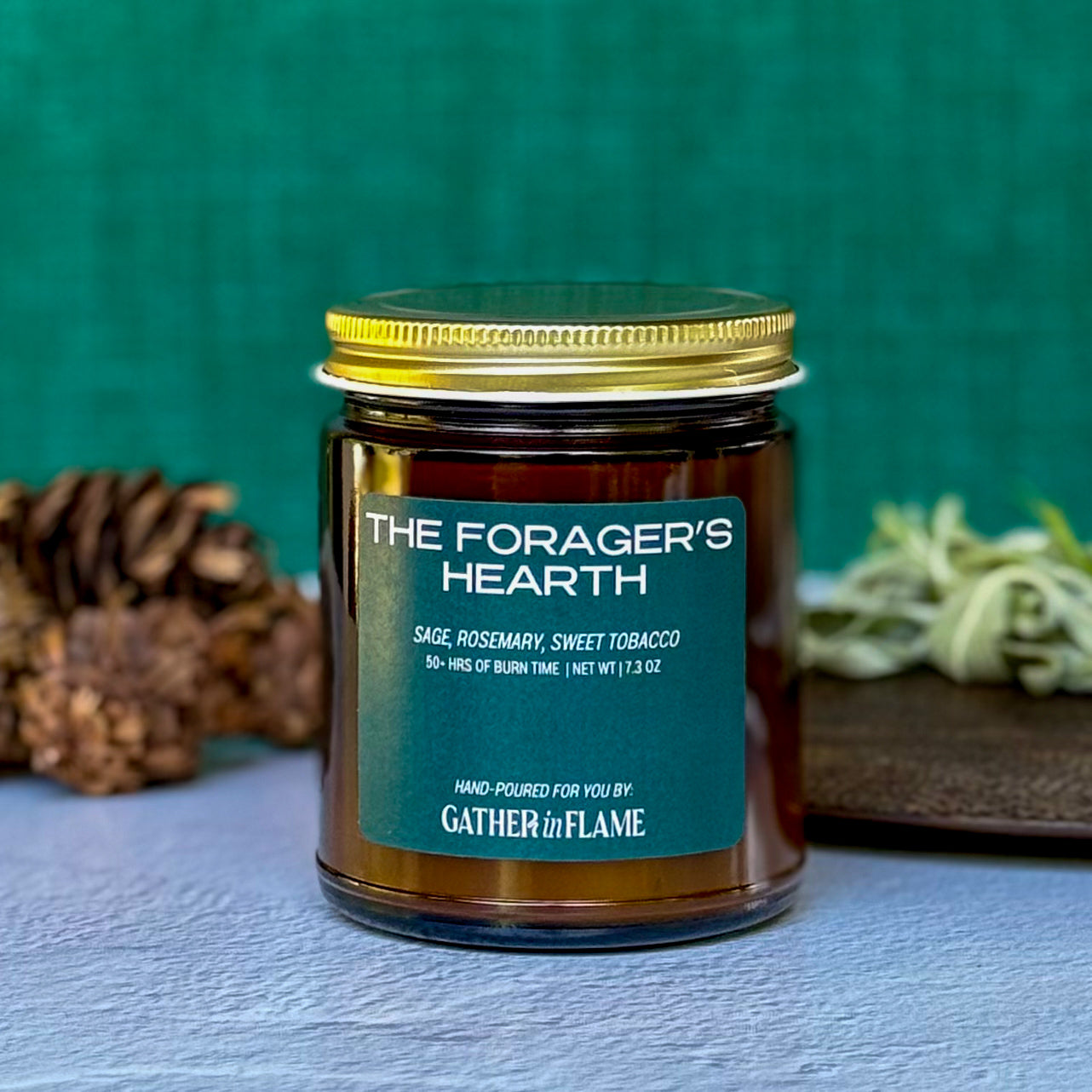 Candle labeled The Forager's Hearth with green background surrounded by pinecones and herbs