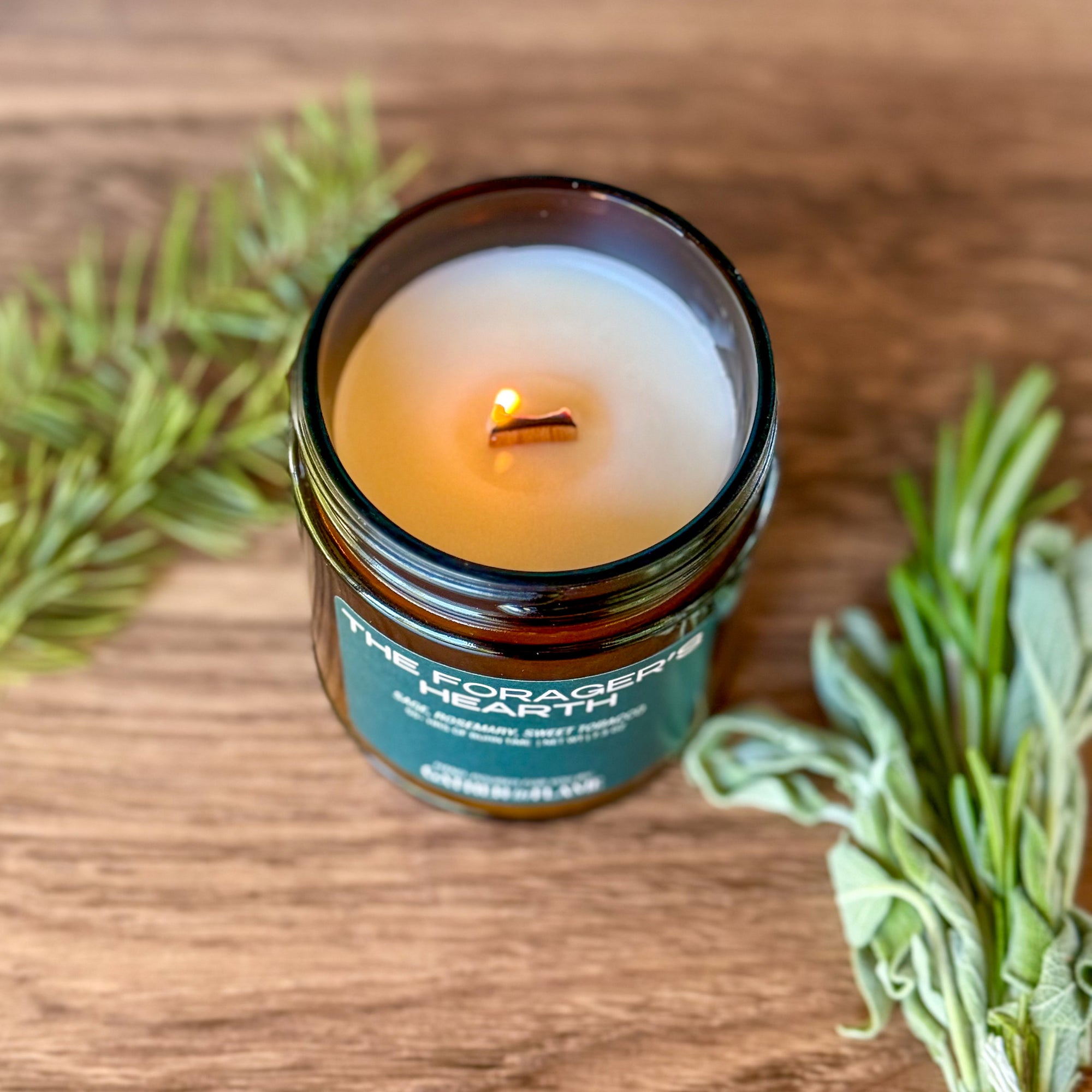 Overhead of lit candle labeled The Forager's Hearth on wooden background surrounded by herbs