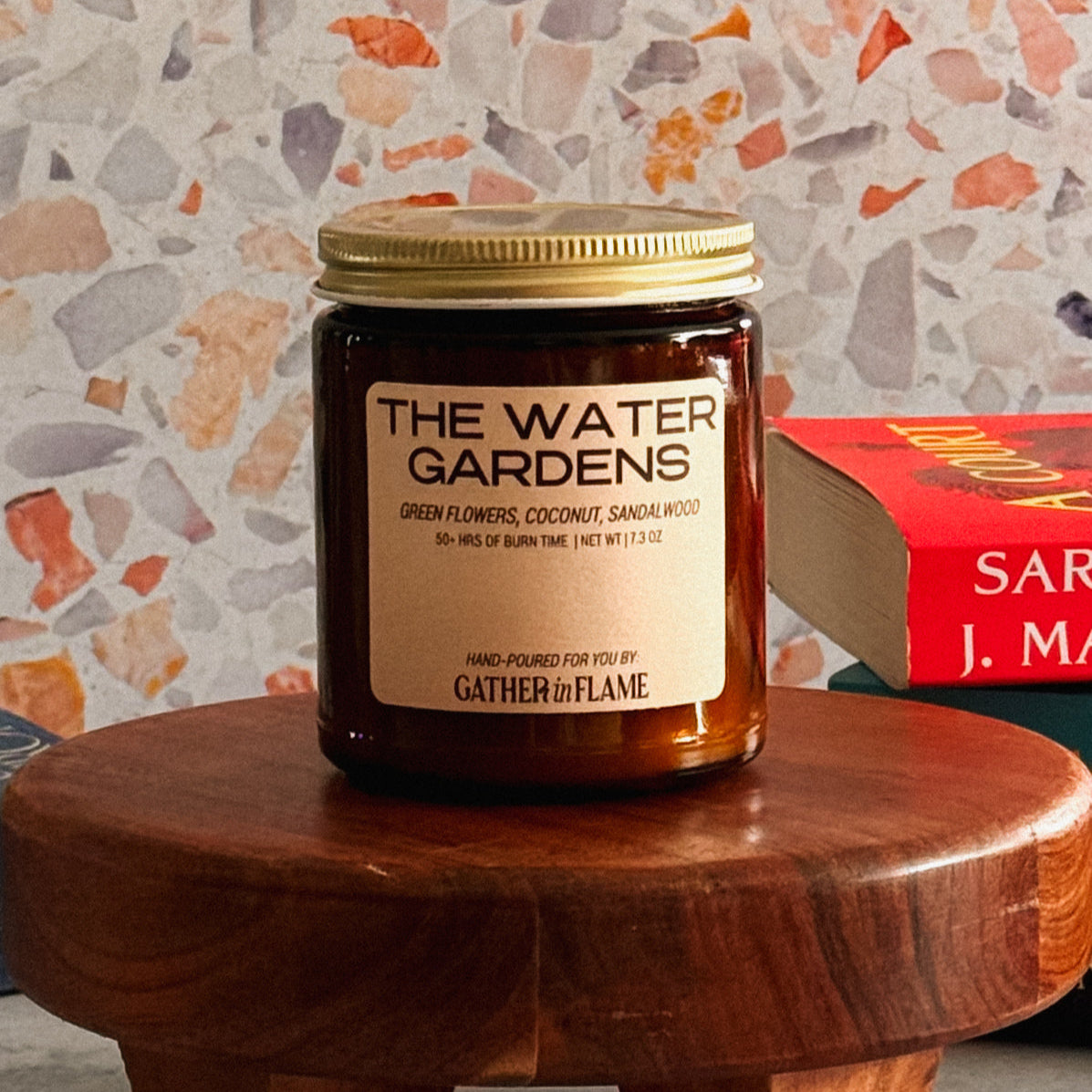 Candle jar labeled 'The Water Gardens' by 'Gather in Flame' on a wooden stool with a patterned wall in the background.