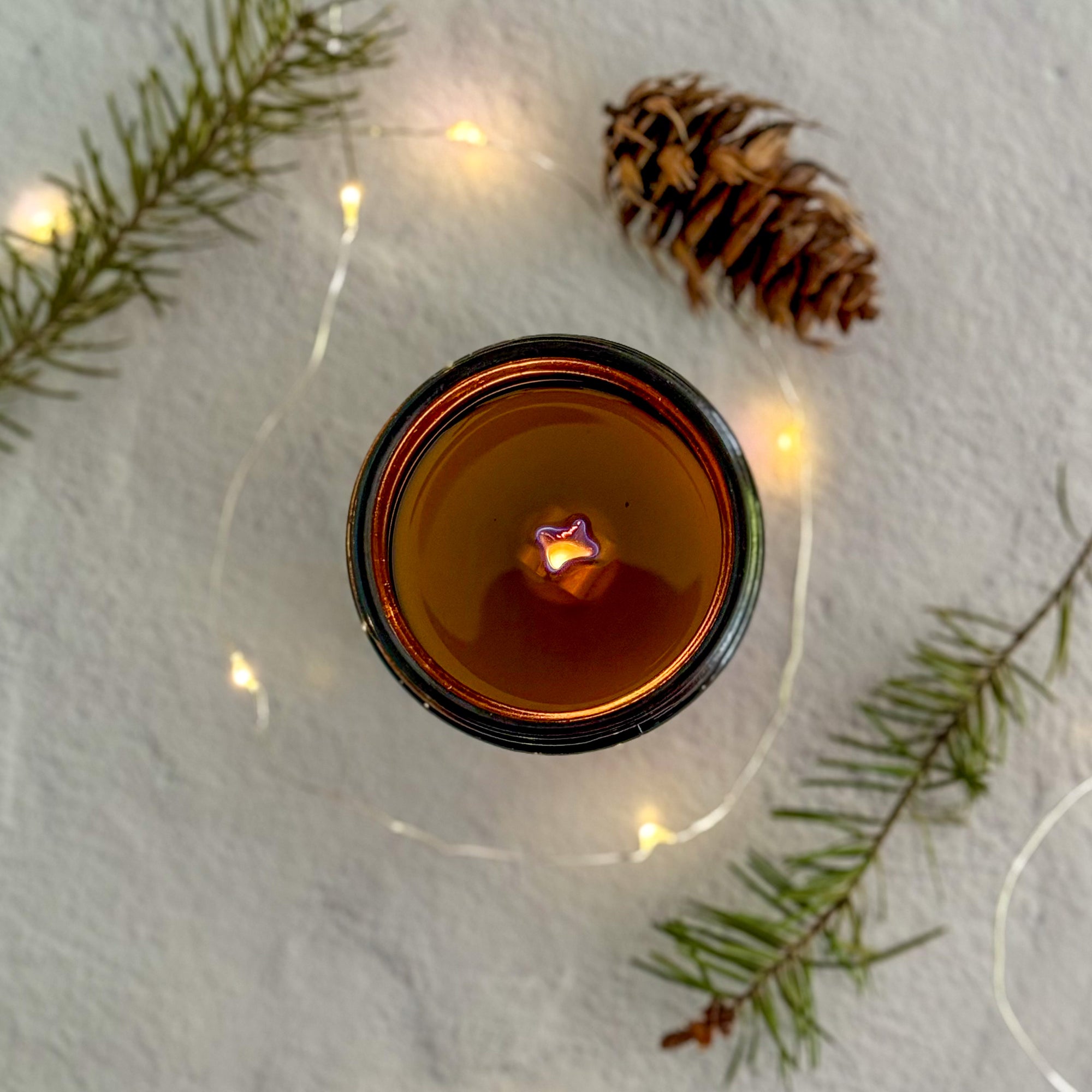 Topdown view of lit candle with wood wick with a pine cone and branches on a textured surface