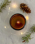 Topdown view of lit candle with wood wick with a pine cone and branches on a textured surface