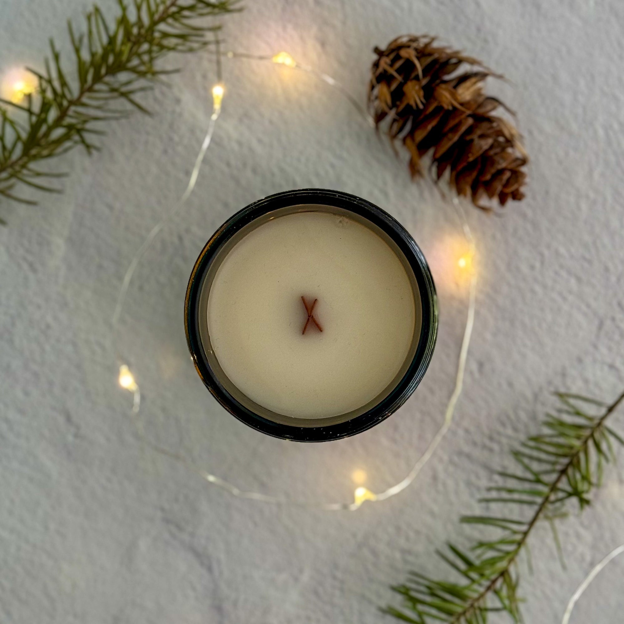 Topdown view of candle with wood wick with a pine cone and branches on a textured surface