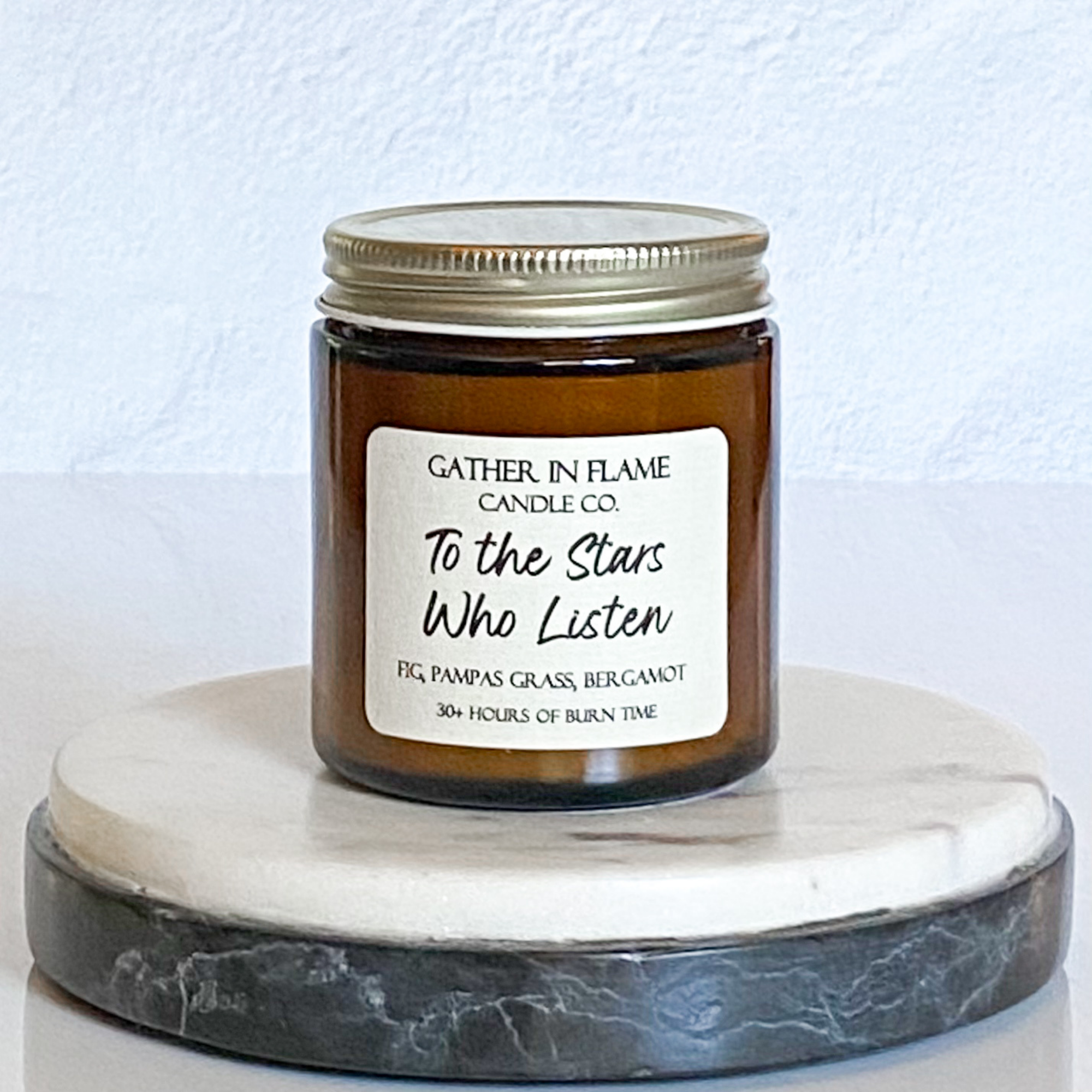 A jar candle with a label that reads 'To The Stars Who Listen' on a white marble coaster. The jar is transparent amber with a gold lid.