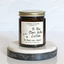A jar candle with a label that reads 'To The Stars Who Listen' on a white marble coaster. The jar is transparent amber with a gold lid.