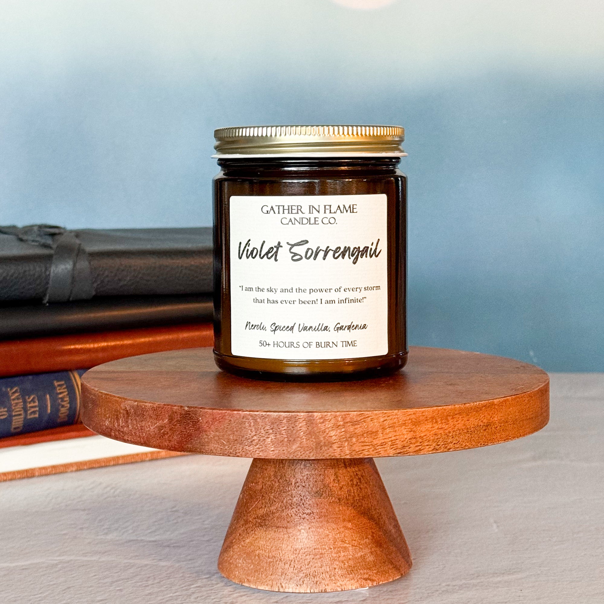 A jar candle with a label reading 'Violet Sorrengail', placed on a wooden stand, with books in the background.