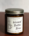Close up of a jar candle with a cream label that reads 'Wizard Butter Brew' with a illustration of a mug, on a brown surface.
