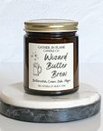A jar candle with a cream label that reads 'Wizard Butter Brew' with a illustration of a mug, on a marble surface.