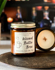 Close up of a jar candle with a cream label that reads 'Wizard Butter Brew' with a illustration of a mug next to candle showing creamy surface and wood x wick, on a wood surface with a plant and bottles in the background.