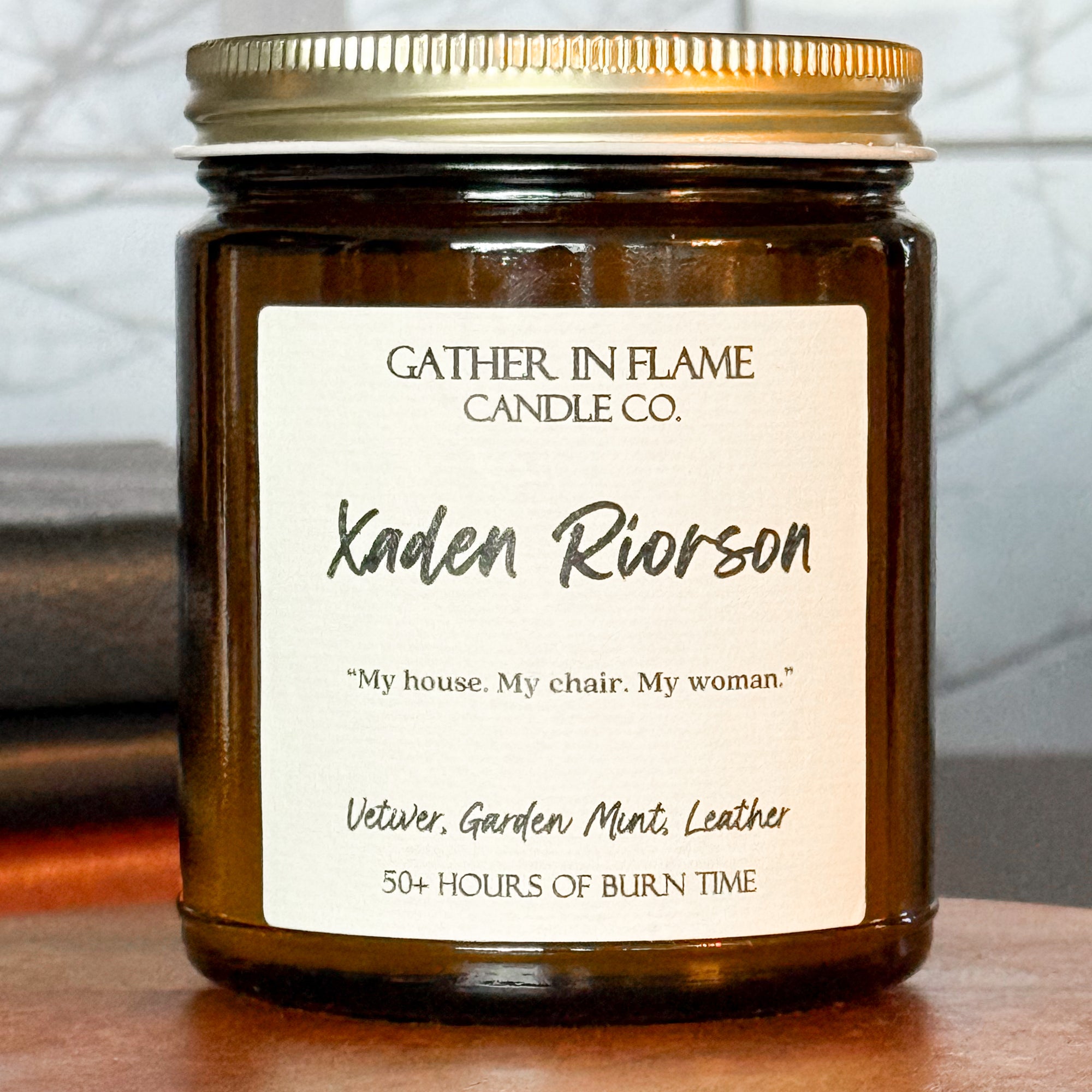 Close up of a jar candle with a gold lid, labeled 'Xaden Riorson', placed on a wooden stand, with books and a window in the background.
