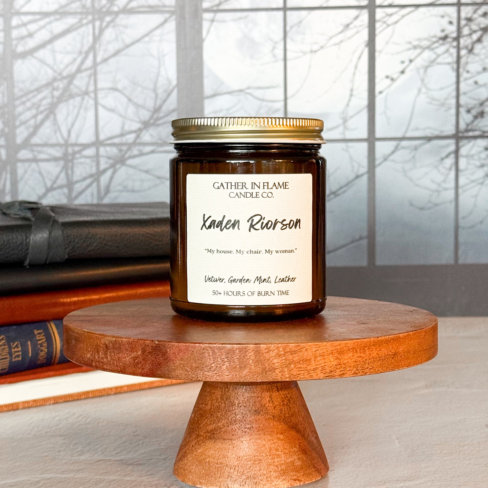 A jar candle with a gold lid, labeled 'Xaden Riorson', placed on a wooden stand, with books and a window in the background.