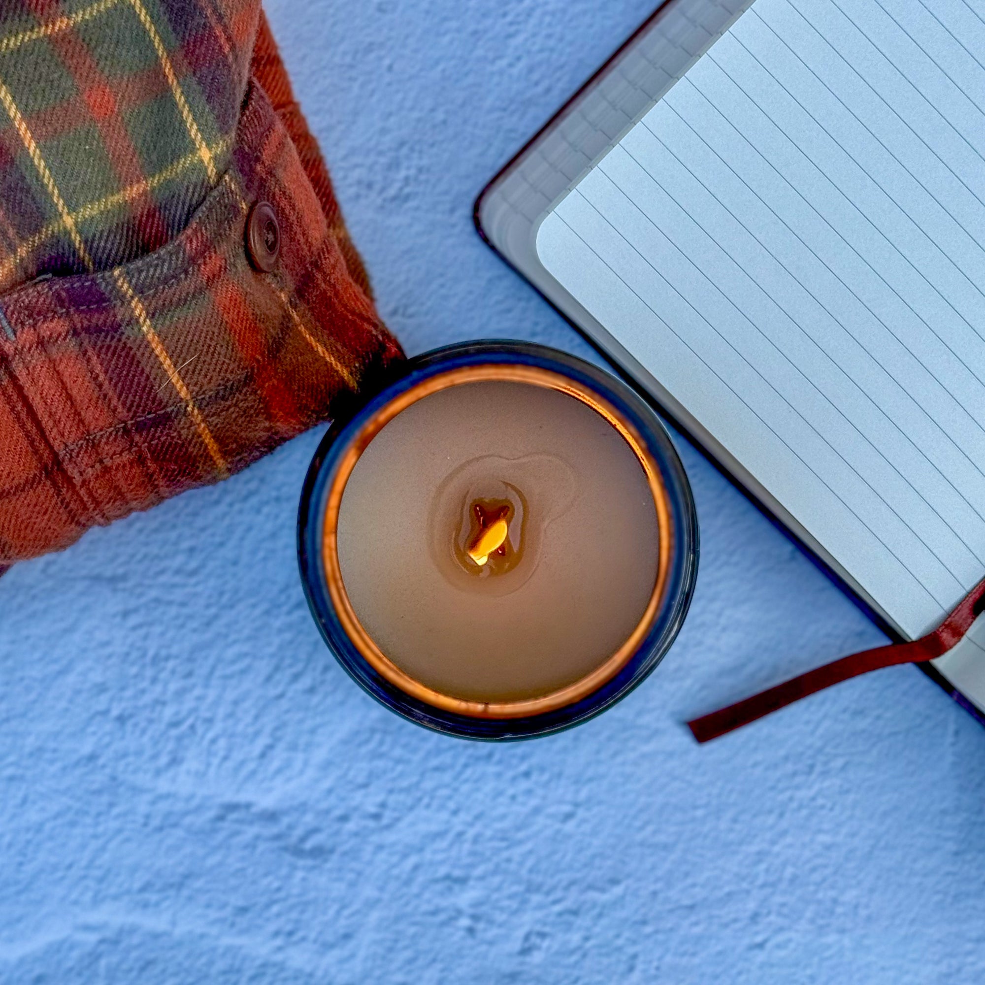 Topdown view of lit candle with wood wick with a notebook and plaid flannel shirt on a blue surface