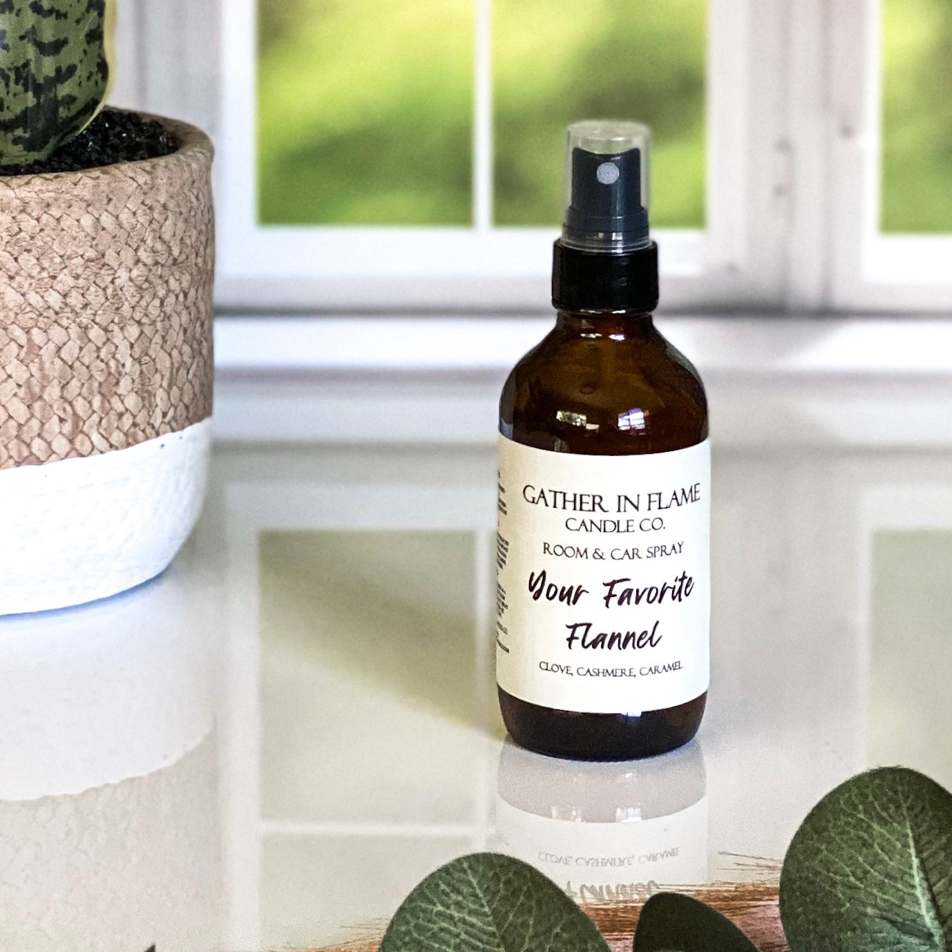 A brown amber spray bottle labeled 'Your Favorite Flannel' by Gather In Flame Candle Co. on a white marble surface with a window and plant in the background.