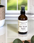 A brown amber spray bottle labeled 'Your Favorite Flannel' by Gather In Flame Candle Co. on a white marble surface with a window and plant in the background.