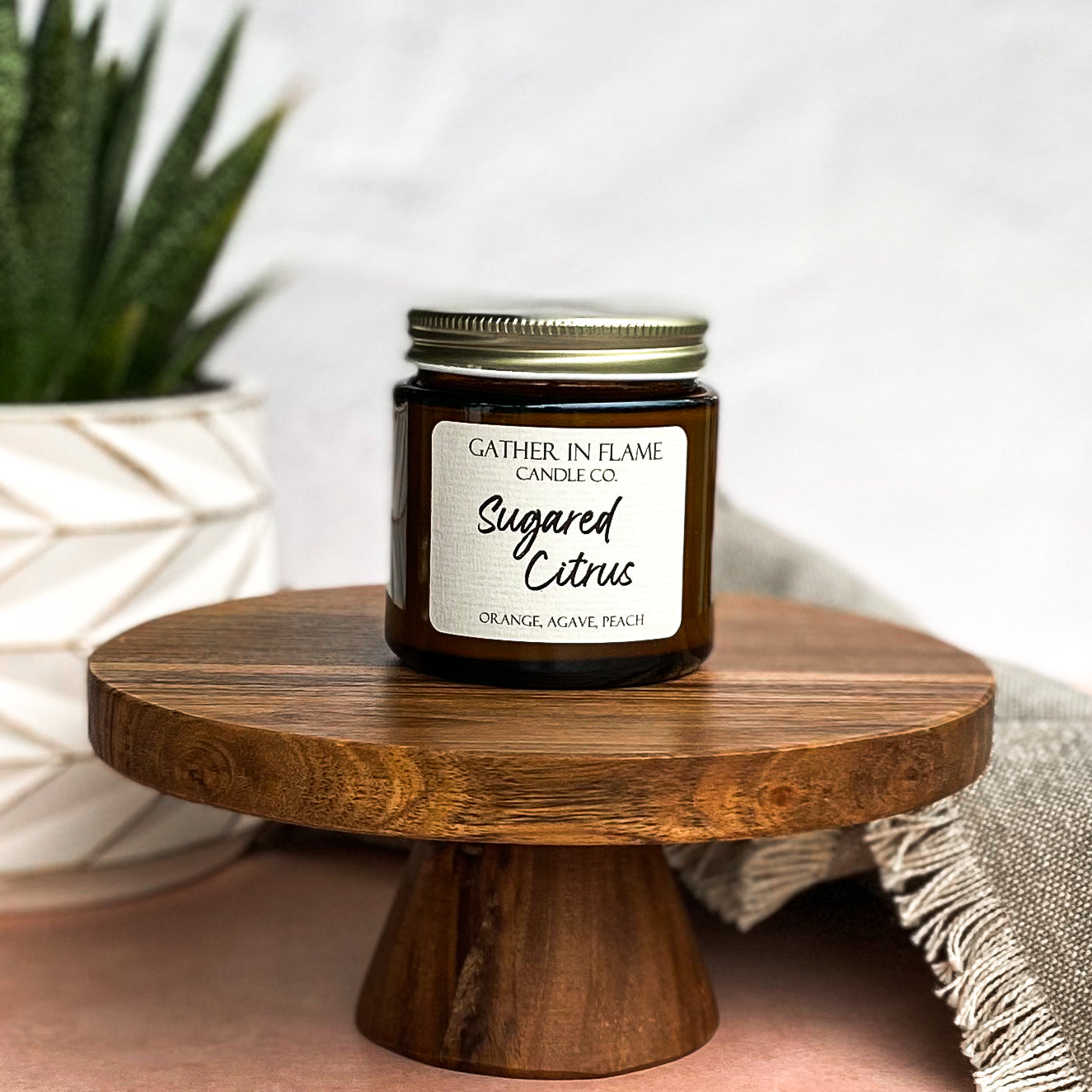 3.6oz candle jar of SSugared Citrus on wooden pedestal next to plant